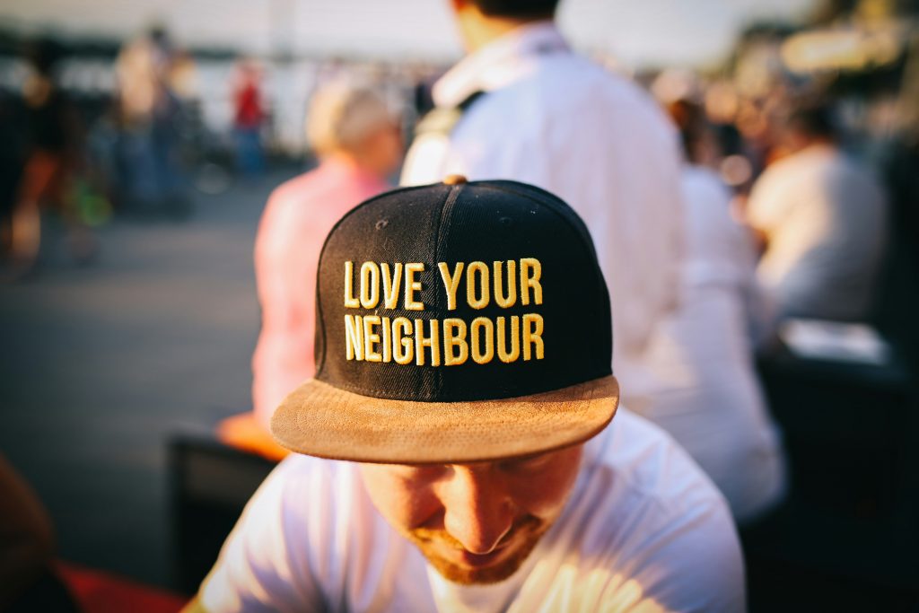 A close-up of a black baseball cap with bold yellow lettering that reads 'LOVE YOUR NEIGHBOUR', worn by a person in a busy outdoor setting, representing kindness, inclusion and community care.