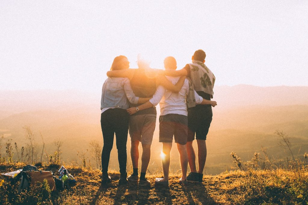 A group of four people standing close together with their arms around each other, facing a golden sunset over a scenic landscape, symbolising community, support and togetherness.
